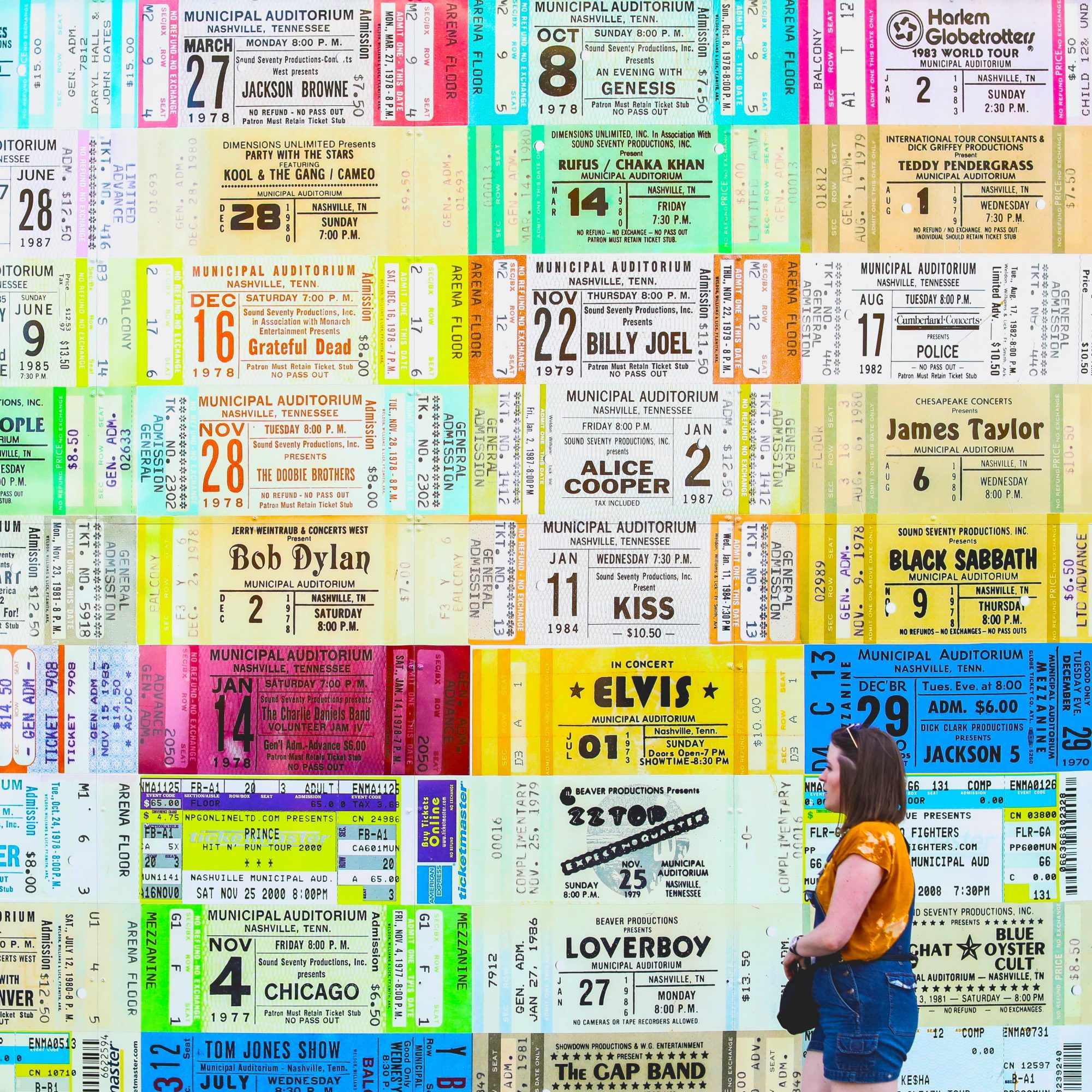 A young woman walks by a wall of tickets. SelfishMe Travel LLC